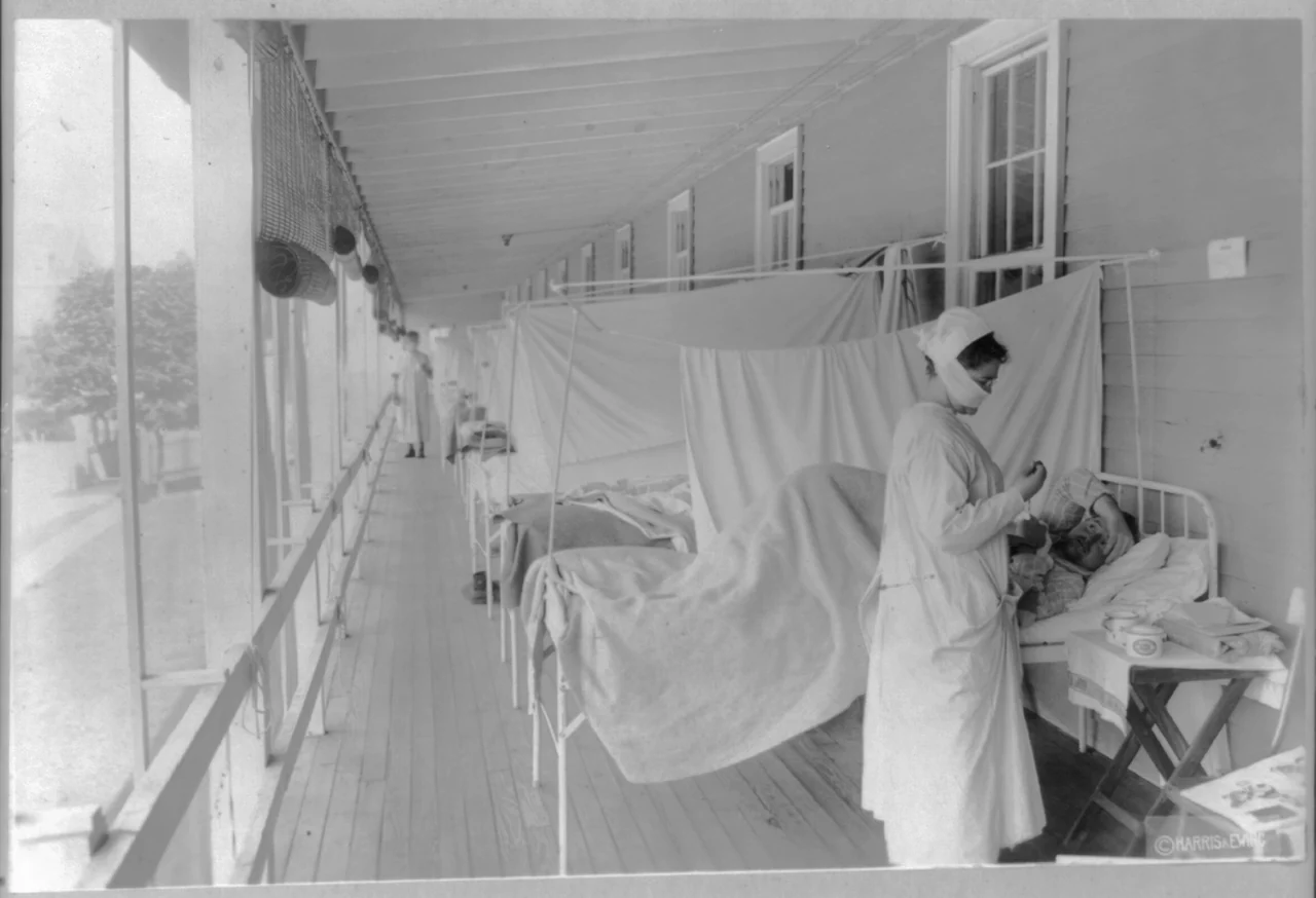 A masked nurse takes a patient’s pulse beside a line of hospital beds on a long open-sided ward.